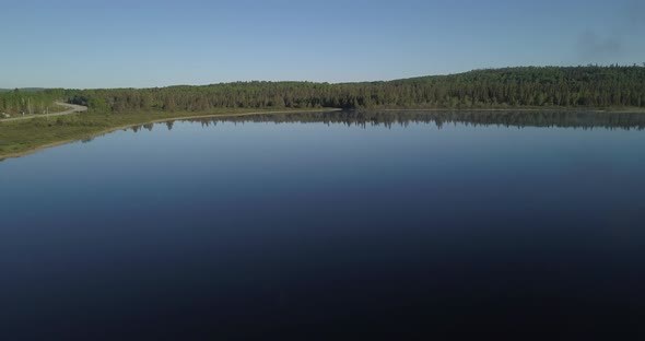 Aerial shot flying over a blue still watered lake facing a beautiful lush green forest in northern O