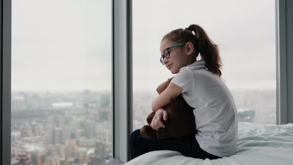 Cute Teenage Girl Sits Hugging Her Favorite Toy on the Bed Near the Window with a City View From