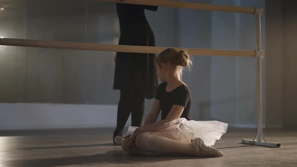 Side View of Inspired Pretty Caucasian Girl in Tutu and Pointes Sitting on Floor Admiring