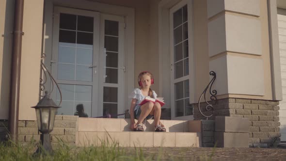 Cute Girl During Summer Vacation Sitting on the Courtyard Near House Listening To Music, Singing