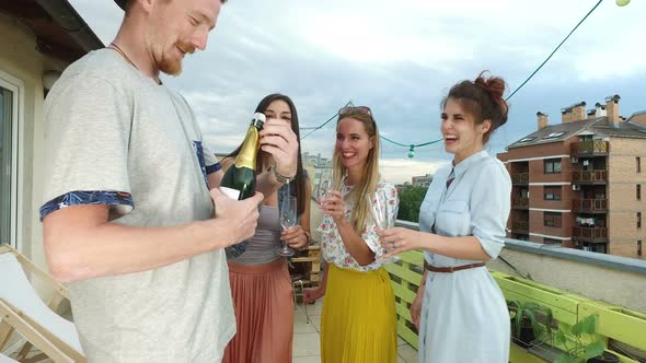 Side view of hipster man opening champagne, female friends waiting with glasses