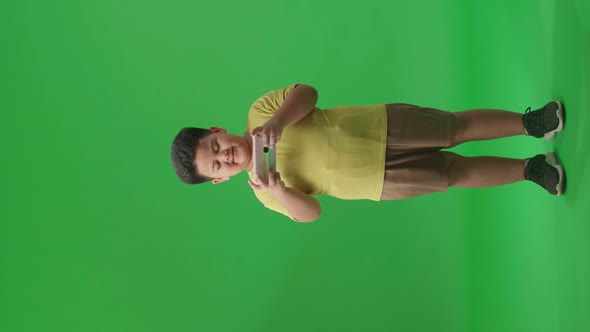 Full Body Of Smiling Asian Little Boy Using Mobile Phone In Green Screen Studio alt