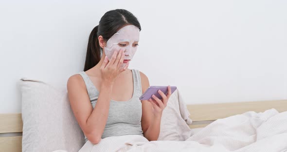 Woman apply facial paper mask on face and use of mobile phone at home