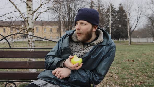 Hungry Bearded Homeless Man Sits on a Bench and Eats a Green Apple in a City Park