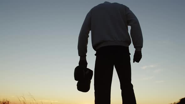 Silhouette man in hoodie walks up hill with boxing gloves in hand and raises hand up