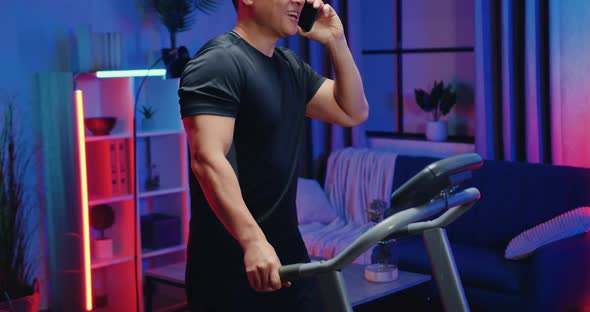 Asian Guy in Training Clothes Walking on Treadmill Simultaneously Talking on Smartphone