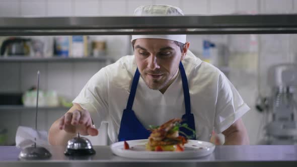 Rack Focus From Confident Smiling Cook Putting Plate with Grilled Prawn Ringing Service Bell to