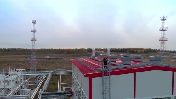 A Drone Flies an Oil and Gas Worker in an Orange Helmet and Overalls Who Stands on the Roof of a Gas
