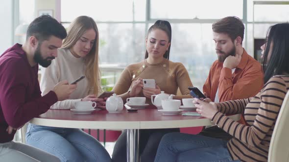 Portrait of Five Bored Caucasian People Sitting in Cafe and Using Smartphones. Young Indifferent Men