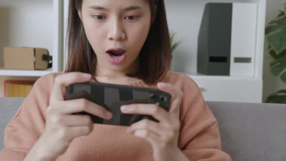 Excited young Asian woman playing games on smartphone in the living room at home.