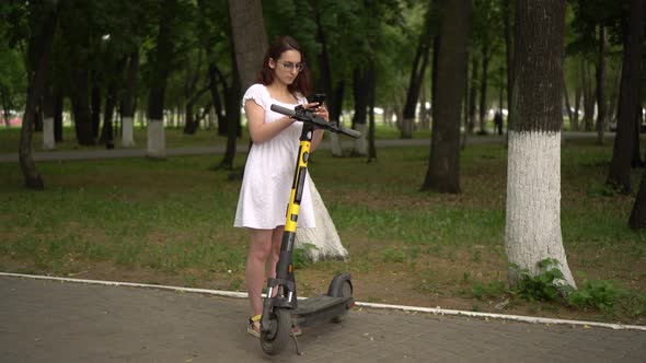 Young Asian Woman in a White Dress Starts a Ride on a Sharing Electro Scooter in the Park