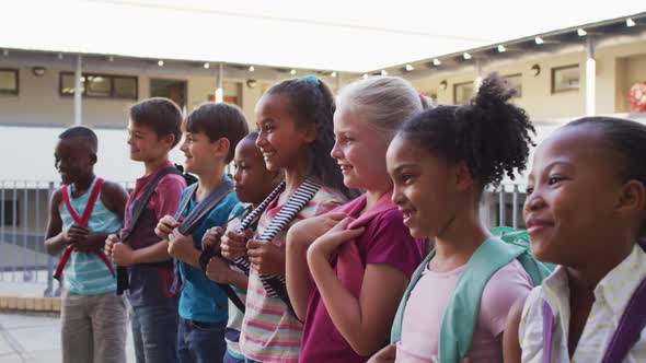 Diverse group of schoolchildren wearing backpacks smiling and standing in a row at school yard