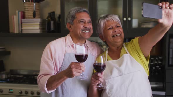 Happy senior mixed race couple taking selfie toasting with wine in kitchen