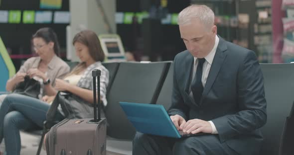 Serious Mid-adult Caucasian Businessman Using Laptop in Airport Waiting Area with People Talking at