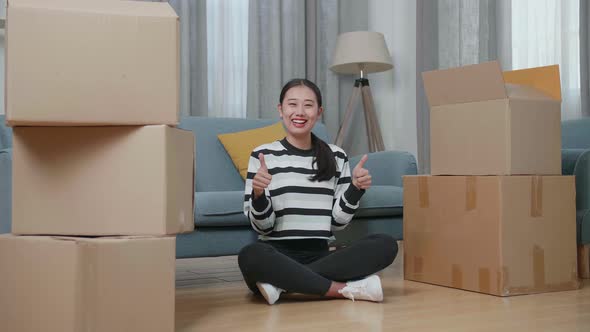 Asian Woman With Cardboard Boxes Smiling And Showing Thumbs Up Gesture To Camera In The New House