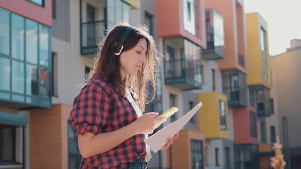 Attractive Business Woman with Documents in Headphones Communicates on a Smartphone with Colleagues