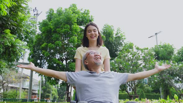 Attractive girl daughter running and pushing elderly father raising arm on wheelchair with smile.