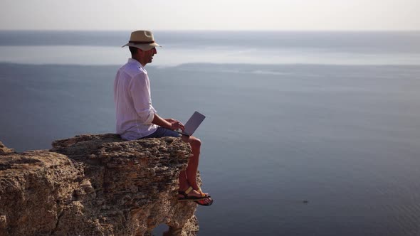 Digital Nomad Man in the Hat a Businessman with a Laptop Sits on the Rocks By the Sea During Sunset