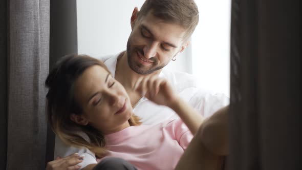 Young Couple Is Sitting By the Window at Home. Husband or Boyfriend Hugs His Wife or Girlfriend