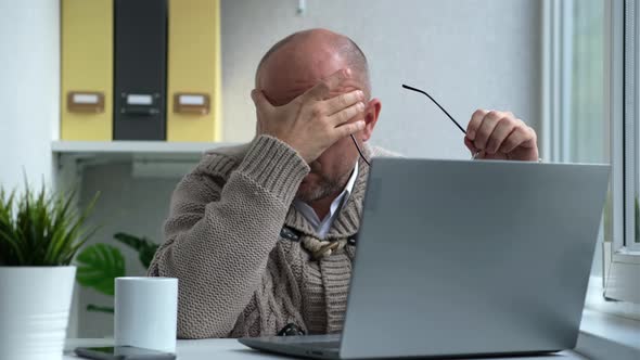 Middleaged Serious Male is Looking at Laptop Taking Off Glasses Tired While Sitting at Table