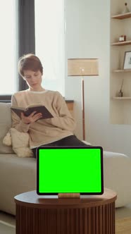 A Woman Reading a Book Sitting on a Sofa in Front of a Green Screen Tablet