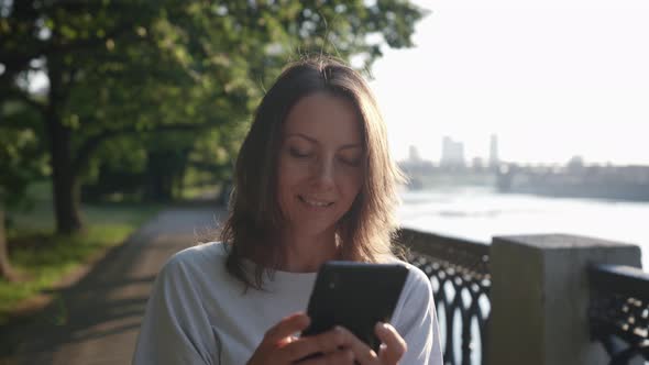 a Woman Carefully Looks at the Phone Screen and Smiles on the City Embankment