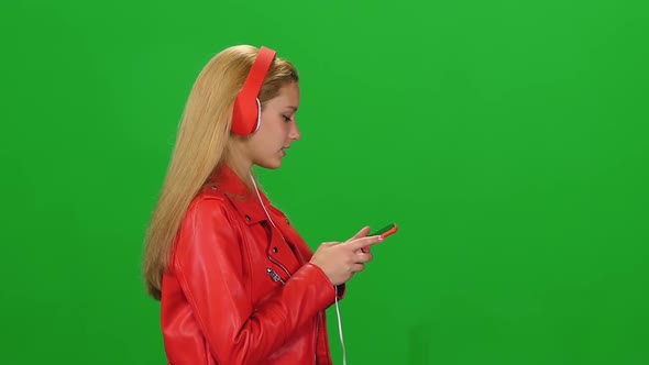 Portrait of Blonde Girl Is Walking in Big Red Headphones, Chooses Music and Moves To Rhythm at Green