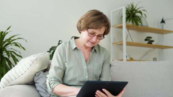 Smiling Elderly Woman Resting on Sofa Using Digital Tablet at Home