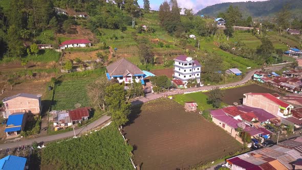 Aerial view of a village in Indonesia surrounded by vegetable farms, trees, fields, volcanoes, and m