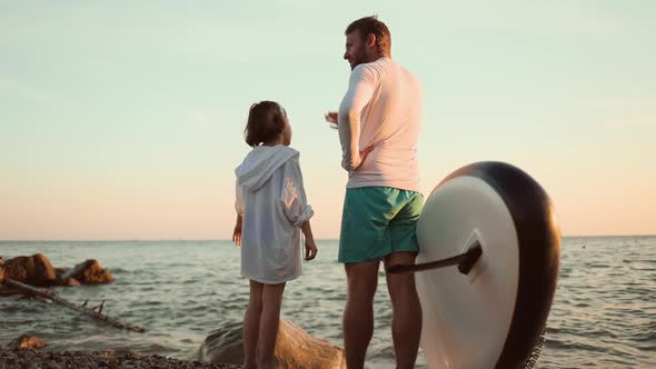 Father and daughter stands on the ocean's beach with a sup board and talking to each other.