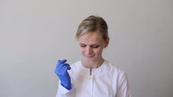 Blonde Female Doctor Surgeon in White Uniform Puts on Blue Gloves and Medical Mask