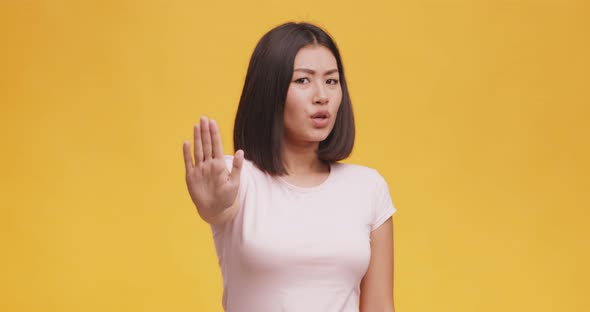 Displeased Young Asian Woman Saying No, Showing Stop Gesture with Palm, Orange Studio Background