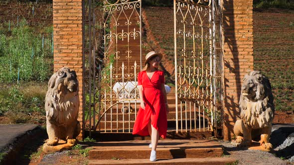 Glamorous Stylish Woman in a Red Dress Walks Near Wrought Iron Gates with Lion Sculptures in a