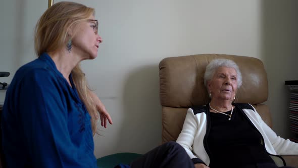 Elderly gray-haired woman sits back in a comfortable chair as she talks to a younger, mature, blonde