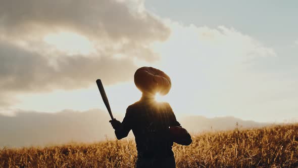 Silhouette of a Man in a Giant Pumpkin Head at Sunset in a Field with a Bat in His Hands Halloween