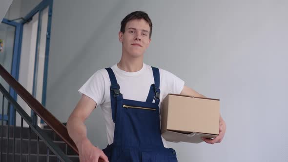 A Courier in a White Tshirt and Blue Overalls Stands on the Stairs in a Highrise Building Holding a