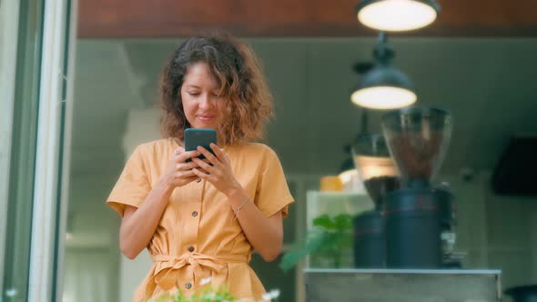 Smiling Curly Woman in Yellow Dress Using Her Phone Happily While Sits in Coffee Shop