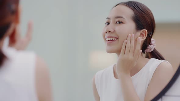 Fresh healthy skin of Asian girl looking at mirror, applying facial moisturizer on her face.