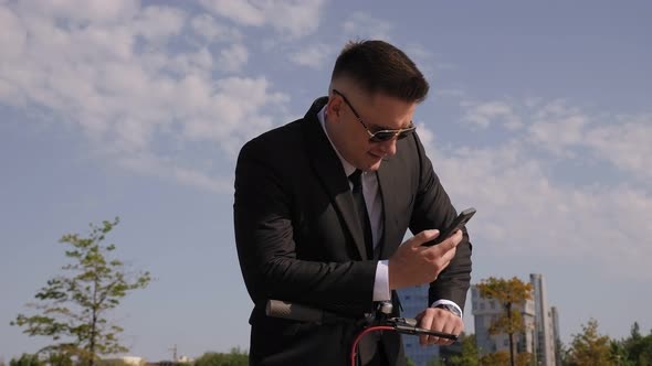 Close-up of a Businessman with a Phone and an Electric Scooter in a Park