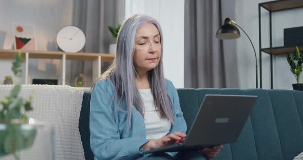 Woman with Long Grey Hair and Casual Clothes Sitting on the Couch at Home and Working on Laptop