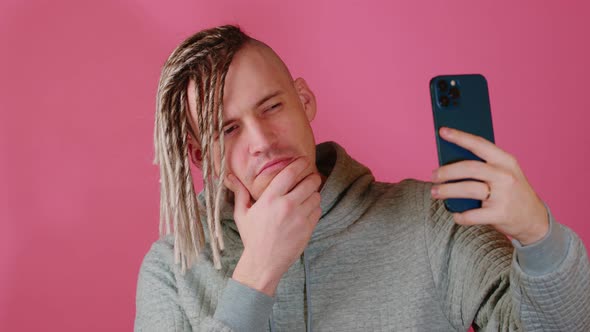 A Handsome White Man with Dreadlocks Takes a Selfie Using a Smartphone on a Pink Background