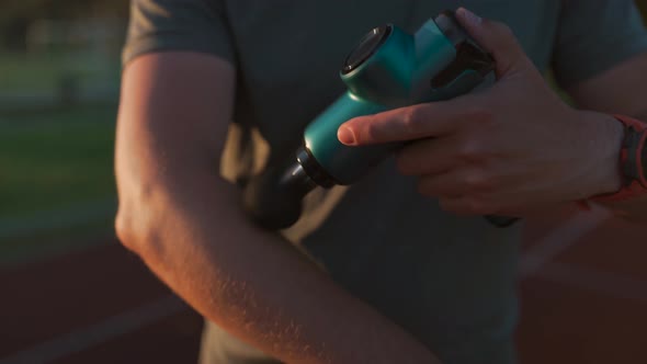 A Male Athlete Massages Muscles and Tendons with a Massage Percussion Device After a Workout at the