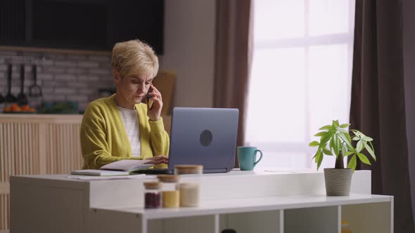 Elderly Woman is Working From Home Sitting at Kitchen and Calling By Cell Phone Using Laptop Medium