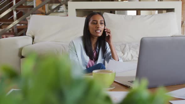 Mixed race woman using laptop talking on phone drinking coffee working from home