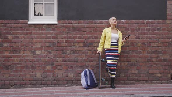 Mixed race woman leaning against a brick wall with her skateboard and her phone