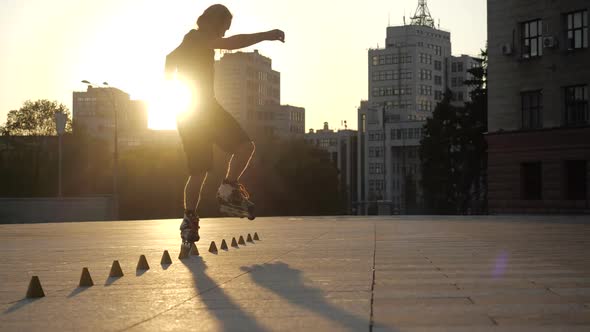 Young Long-haired Man Roller Skater Is Dancing Between Cones in the Evening in a City Square at