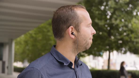 A Caucasian Man Listens to Music with Earphones on By an Office Building in an Urban Area  Closeup