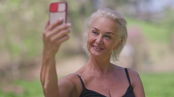 Cheerful Confident Senior Woman Talking Using Video Chat on Smartphone Standing in Sunny Summer Park