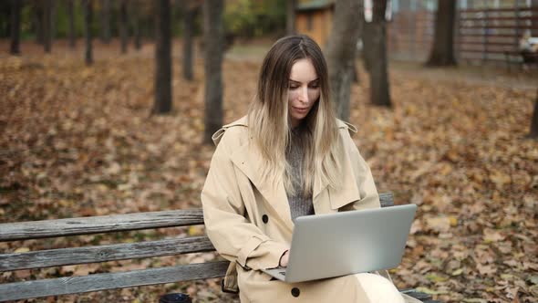 Young Beautiful Woman of European Appearance Works at a Laptop Sitting on a Park Bench