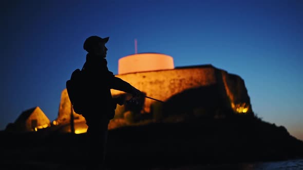 Silhouette of a man fishing with Fort Grey illuminated in the background, at dusk